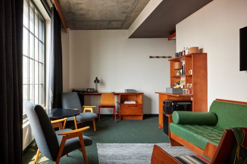 A modern hotel room featuring a green sofa, wooden furniture, a desk, and large windows with natural light.