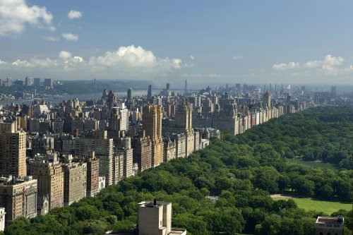 Aerial view of a city skyline with green parks, featuring tall buildings and a river in the background under a clear sky.