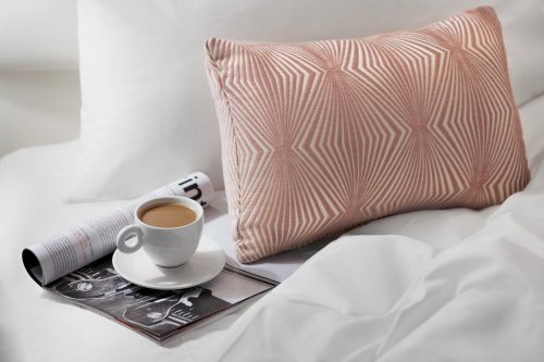A cozy bed scene featuring a patterned pillow, a cup of coffee on a saucer, and a magazine on white bedding.
