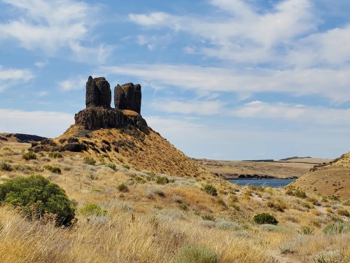 Two tall rock formations atop a grassy hill, with a calm lake and blue sky in the background.