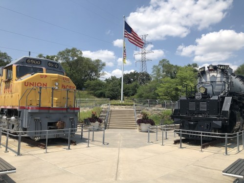 Two historic trains on display, one yellow Union Pacific locomotive and one black steam engine, with an American flag in the background.