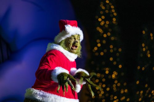 The Grinch in a Santa costume, wearing a red hat and white fur trim, stands against a festive backdrop with lights.