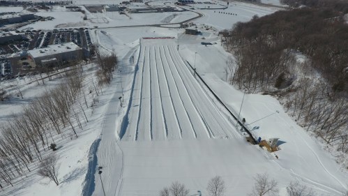 Aerial view of a snowy slope with multiple lanes for tubing, surrounded by trees and a parking lot in the background.