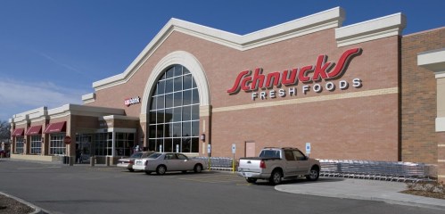 Exterior of a Schnucks Fresh Foods grocery store with large windows and parked cars in front. Clear blue sky above.