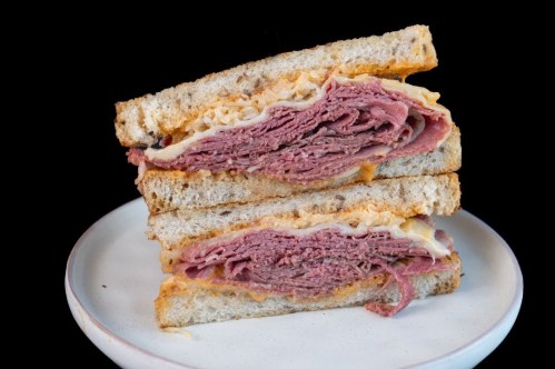 A stacked sandwich with layers of pastrami and cheese on toasted bread, served on a plate against a black background.