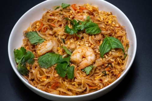 A bowl of pad thai with shrimp, vegetables, and fresh herbs, served on a dark background.