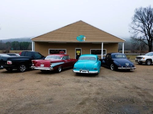 A row of classic cars parked in front of a small, rustic building on a foggy day.
