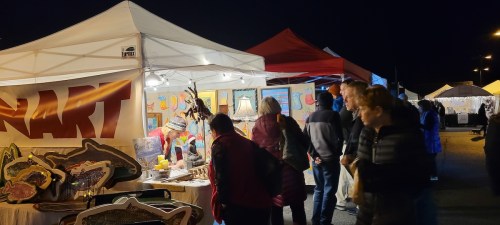 A night market scene with people browsing art stalls under colorful tents, illuminated by warm lights.