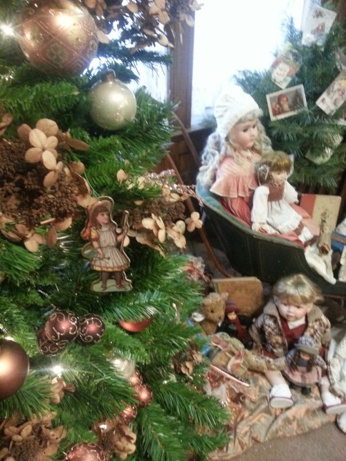 A decorated Christmas tree with ornaments, surrounded by vintage dolls and festive decorations in a cozy setting.