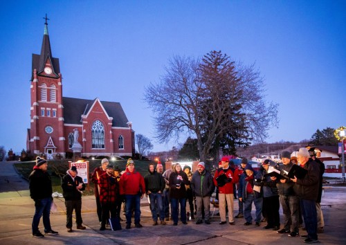 A group of carolers in winter attire sings outside a church at dusk, with snow on the ground and trees nearby.