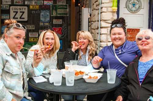 Five women enjoying food at an outdoor table, smiling and holding up pieces of fried food.