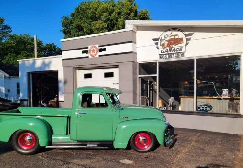 A vintage green pickup truck parked in front of a garage with a sign that says "Open." Trees in the background.
