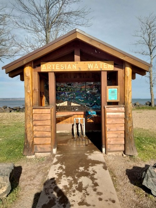 A wooden structure labeled "Artesian Water" with water spouts, surrounded by grass and trees near a body of water.