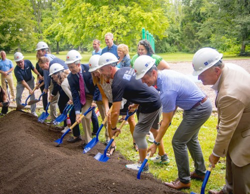 A group of people in hard hats using shovels to break ground at a park ceremony. Trees and playground equipment in the background.