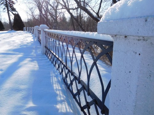 A snow-covered fence stretches along a path, surrounded by trees in a winter landscape.