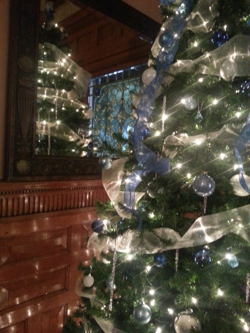 A decorated Christmas tree with blue and silver ornaments, reflecting in a nearby ornate mirror.