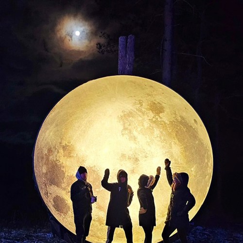 Four people pose in front of a large illuminated moon sculpture at night, with a real moon visible in the sky above.