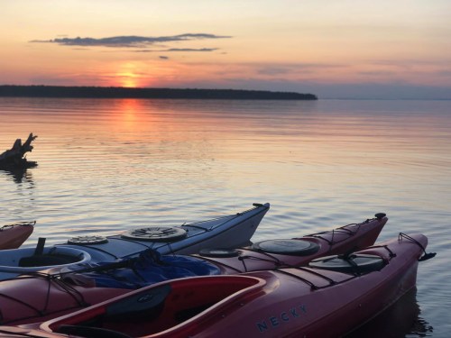 Kayaks on calm water at sunset, with vibrant orange and purple hues reflecting on the surface.