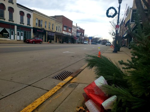 A quiet street lined with shops, decorated for the holidays, featuring a wreath and festive greenery in the foreground.