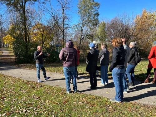 A group of people listens to a speaker in a park on a sunny autumn day, surrounded by trees and fallen leaves.