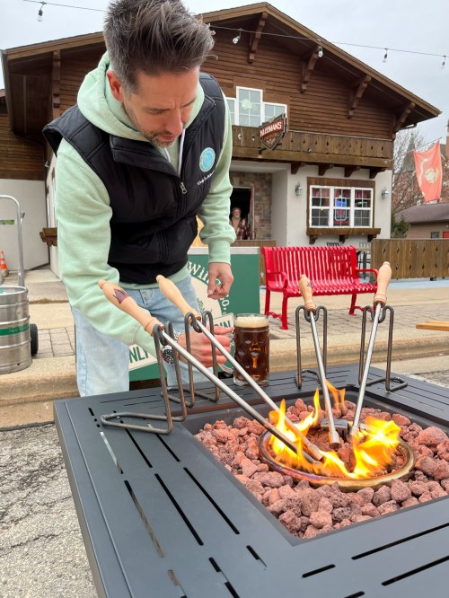 A man in a vest roasts marshmallows over a fire pit, with a beer nearby and a rustic building in the background.