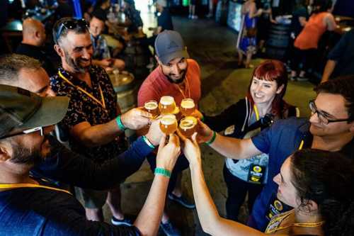 A group of six people cheer with small beer glasses in a lively brewery setting.