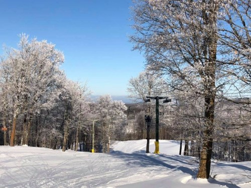 A snowy landscape with frosted trees and a clear blue sky, featuring ski lifts in the background.