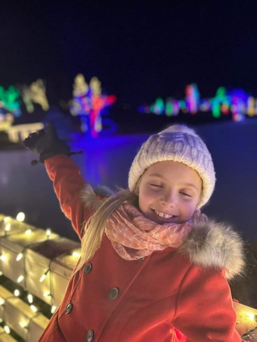 A smiling girl in a red coat and white hat poses joyfully in front of colorful holiday lights at night.