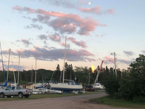 A marina at sunset with sailboats, clouds, and a rising moon in the background. Trees line the horizon.