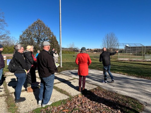 A group of people listens as a man gestures while speaking outdoors on a sunny day.