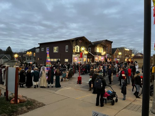 A festive crowd gathers in a town square, with holiday decorations and lights on buildings, under a cloudy sky.
