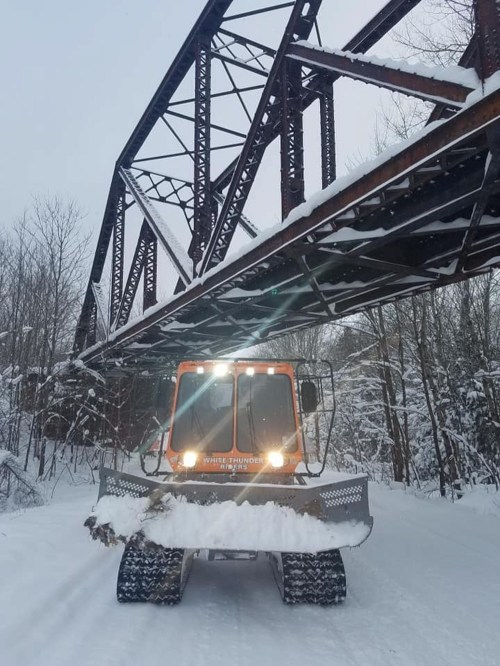 A snowplow clears a snowy path beneath a rusted metal bridge in a winter landscape.