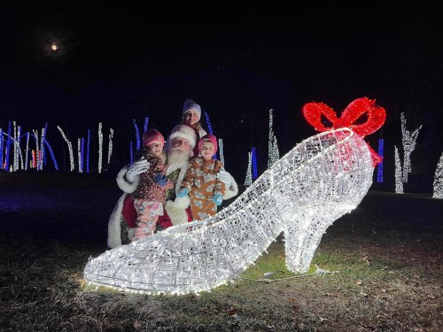 A family poses with Santa next to a large illuminated glass slipper in a festive outdoor setting.