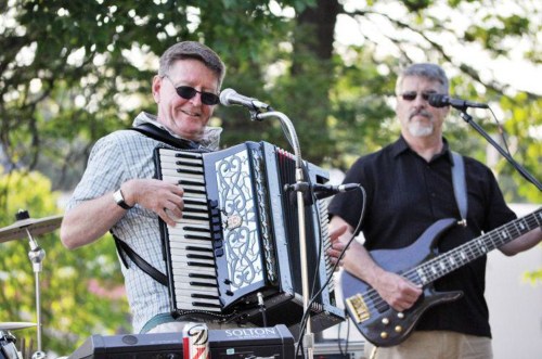 Two musicians perform outdoors; one plays the accordion while the other plays a bass guitar.