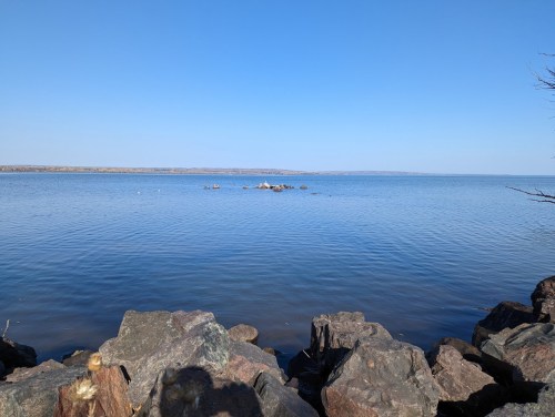 A calm blue lake stretches to the horizon, with rocky shorelines and clear skies above.