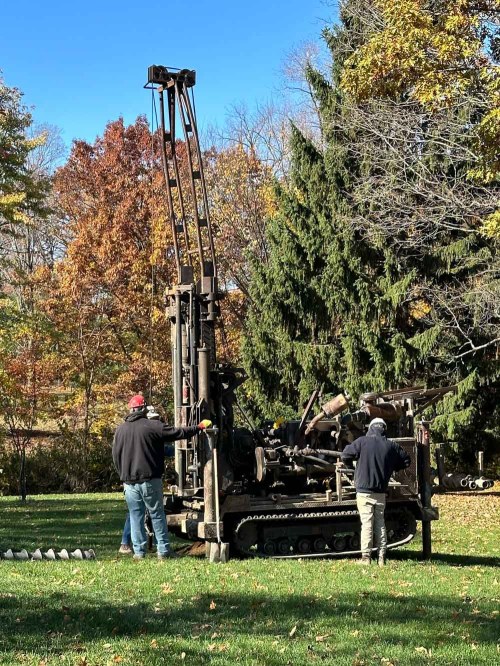 Two workers operate a large drilling machine in a wooded area with autumn foliage.