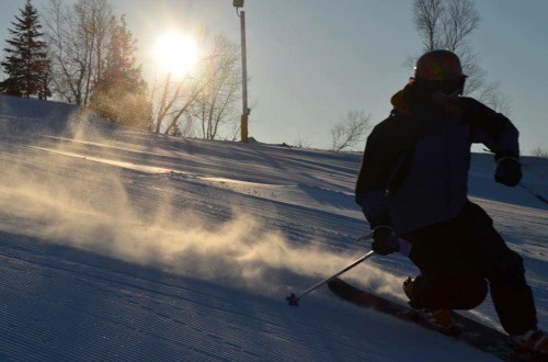 A skier carves through fresh snow with sunlight shining in the background, creating a dynamic winter scene.