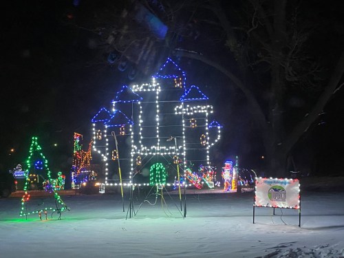 A brightly lit castle made of lights stands in the snow, surrounded by colorful holiday decorations at night.