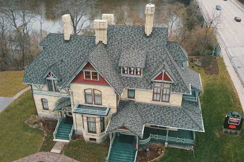 Aerial view of a large, historic mansion with a steep roof, multiple chimneys, and green accents, near a river.