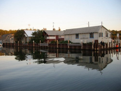 A calm waterfront scene with wooden buildings reflecting in the water at sunset.