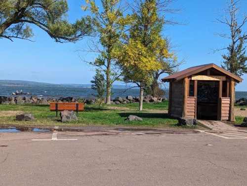 A wooden shelter and bench in a park with trees and a view of a lake in the background.