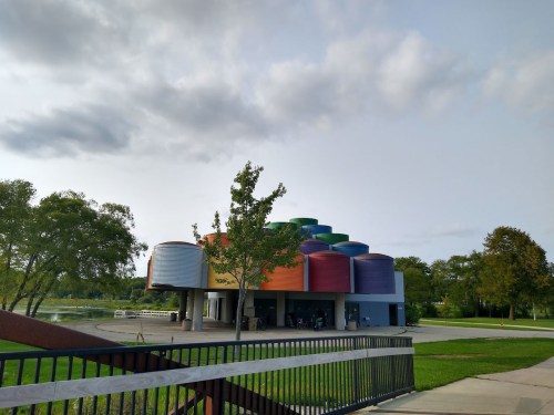 Colorful, modern building with a curved roof, surrounded by trees and a pathway near a body of water under a cloudy sky.