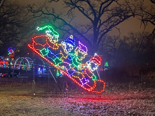 Colorful holiday lights depict three figures sledding down a slope, surrounded by trees in a festive nighttime setting.