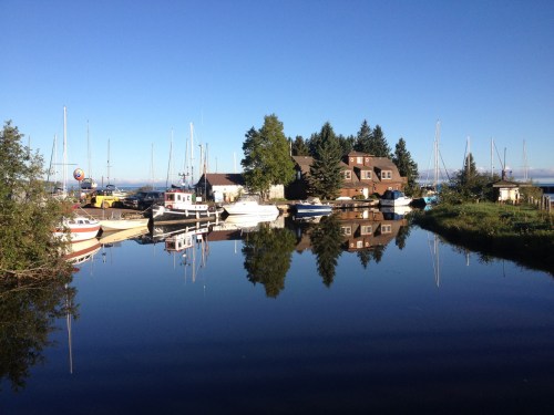 A calm marina scene with boats and a house, reflecting in still water under a clear blue sky.