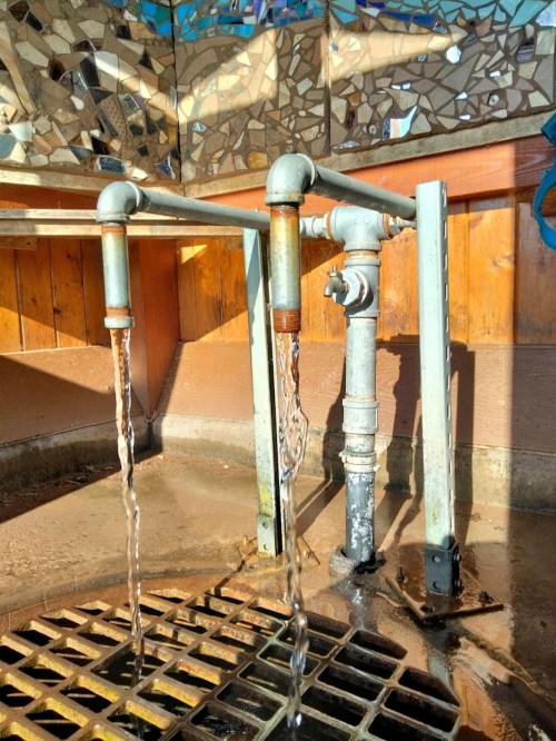 Three metal pipes with water flowing from them, set against a wooden wall and a colorful mosaic background.