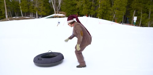 A person in a Santa hat prepares to ride a snow tube on a snowy hill surrounded by trees.