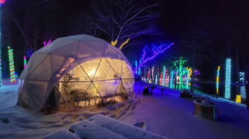 A cozy geodesic dome illuminated with warm lights, surrounded by colorful holiday lights in a snowy landscape at night.