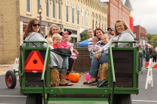 A group of adults and children sitting on a hay-filled wagon during a festive parade.