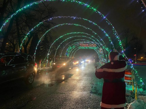 A festive light display arches over a road, with cars passing and a person in a Santa suit waving.