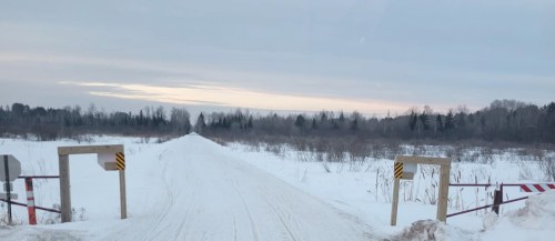 Snow-covered road leading into a winter landscape, flanked by barriers and trees under a cloudy sky.
